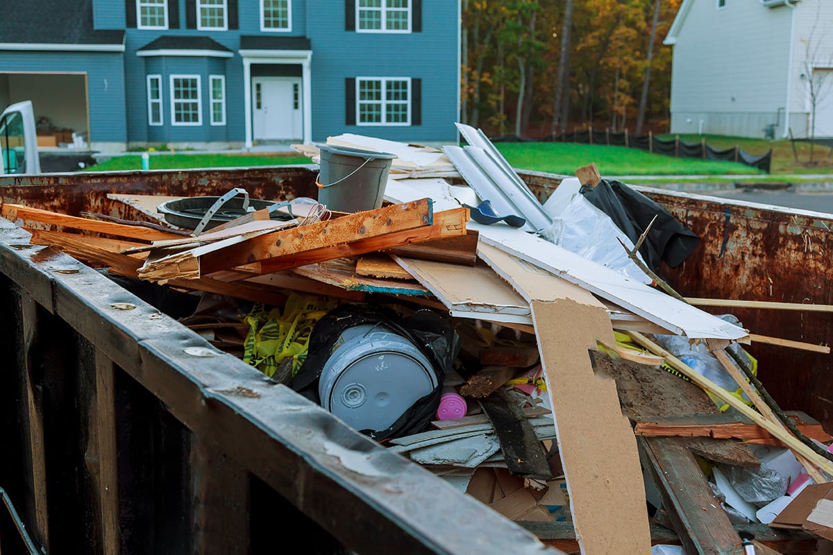 Wide-angle view of an open construction dumpster filled with mixed debris in a residential neighborhood with new home construction in the background.