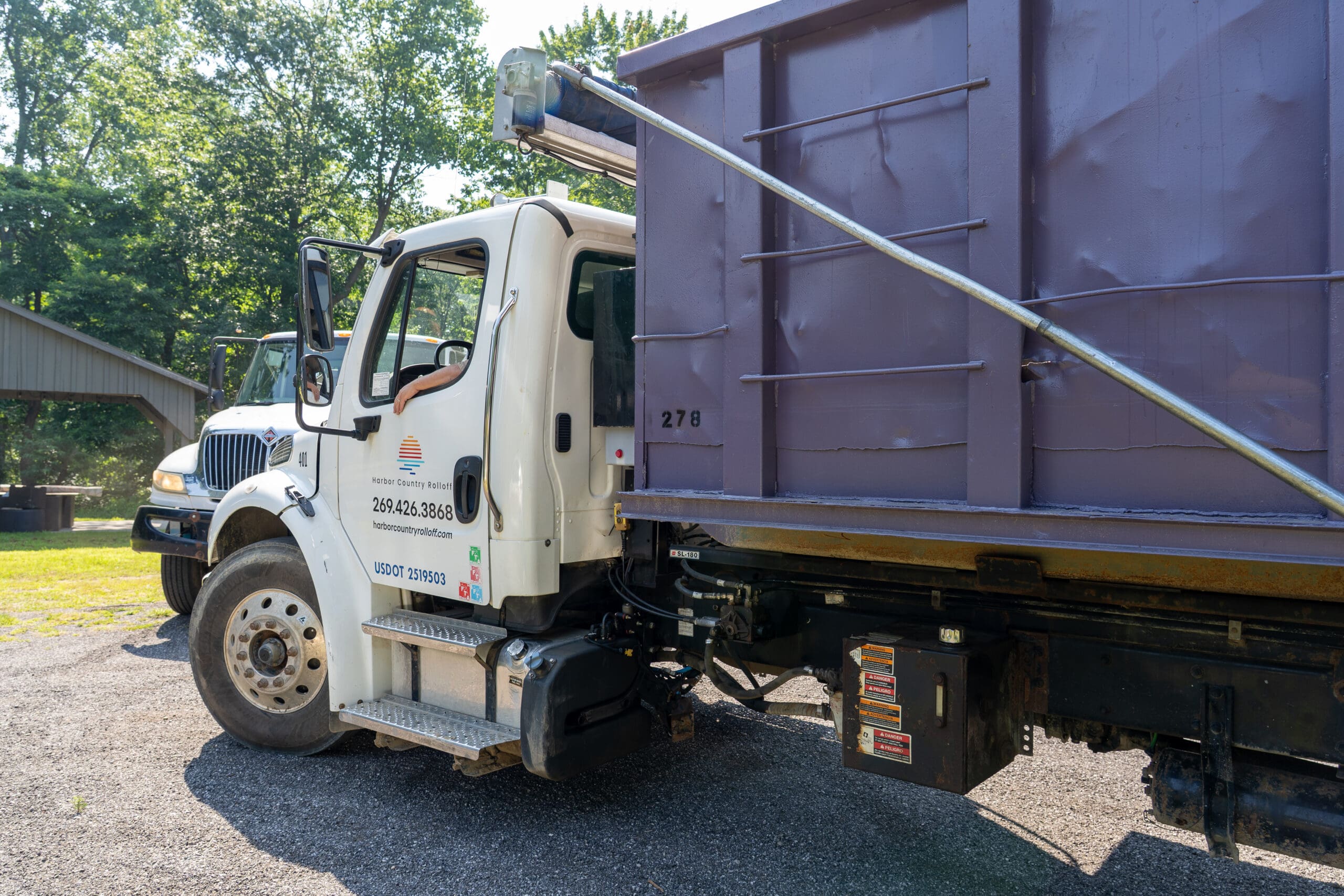 Side view of Harbor Country Rolloff truck with a purple dumpster secured on the bed and the driver’s arm visible through the window.