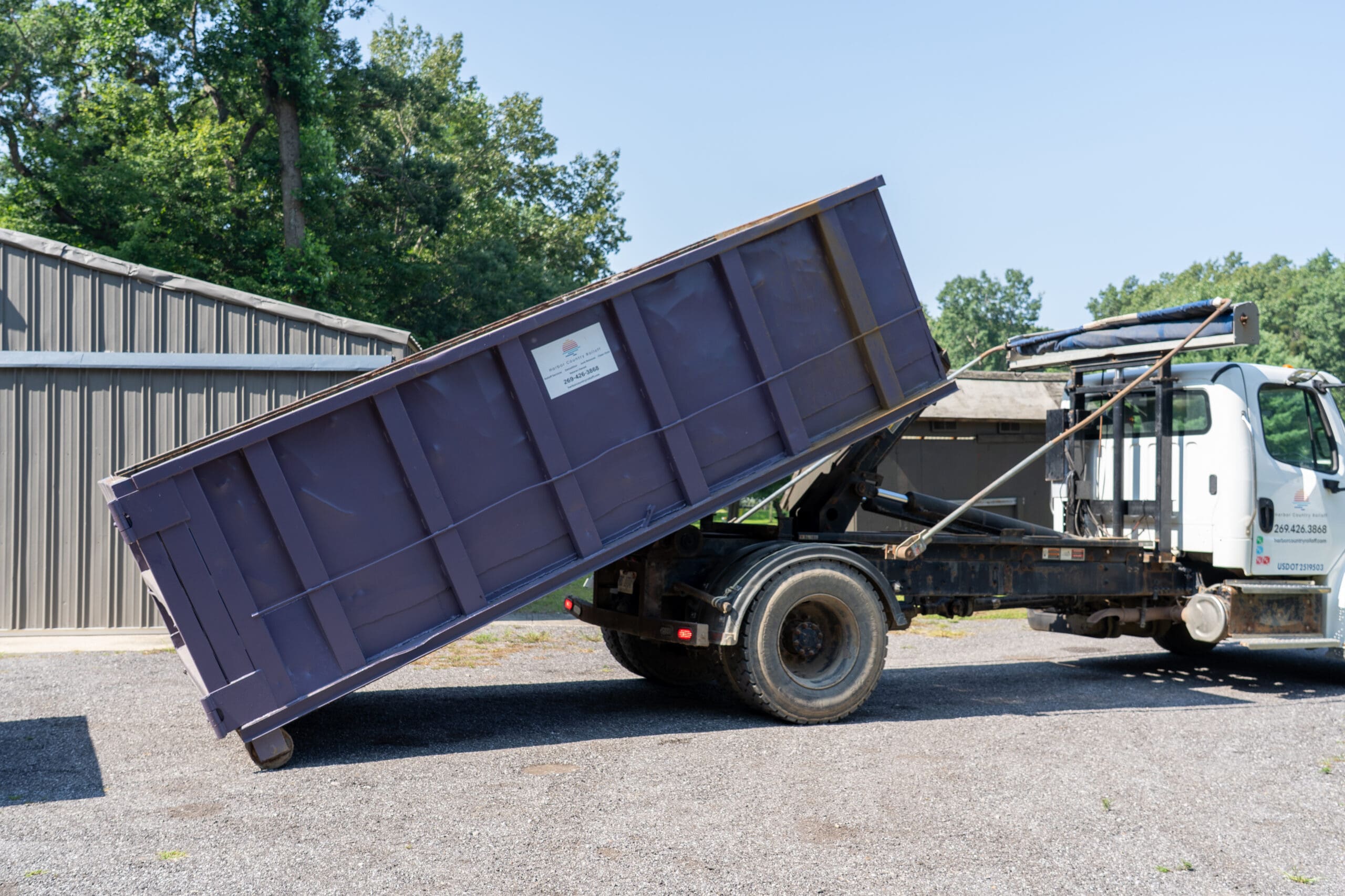 Harbor Country Rolloff truck unloading a purple dumpster on gravel at a job site near a metal building.