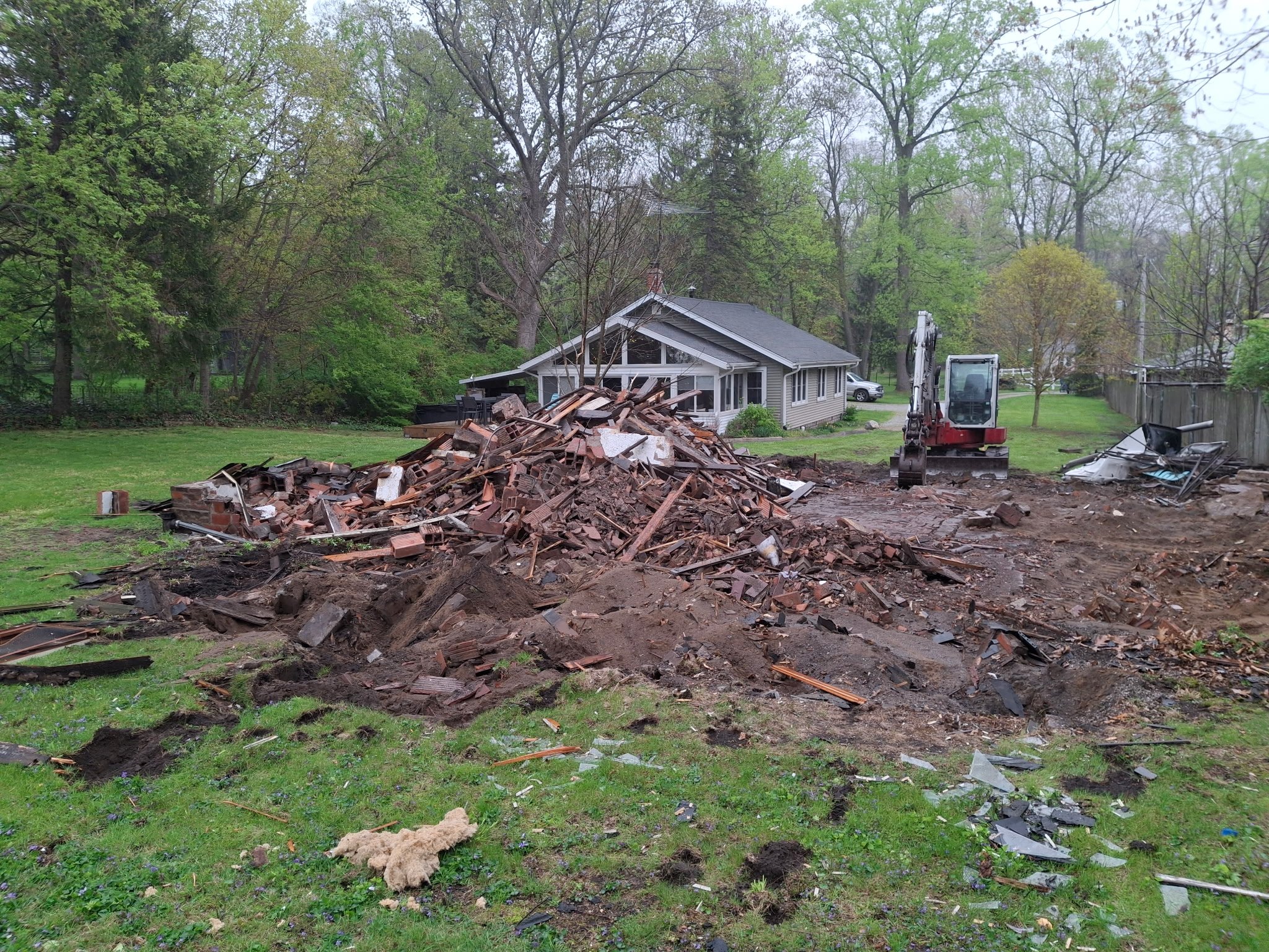 Demolition debris piled on a residential lawn after tear-down, with an excavator on-site for debris removal and site clearing.